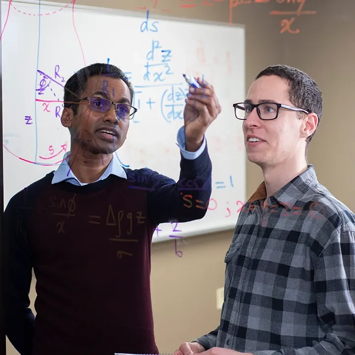 Two male students performing math equations on a glass wall.