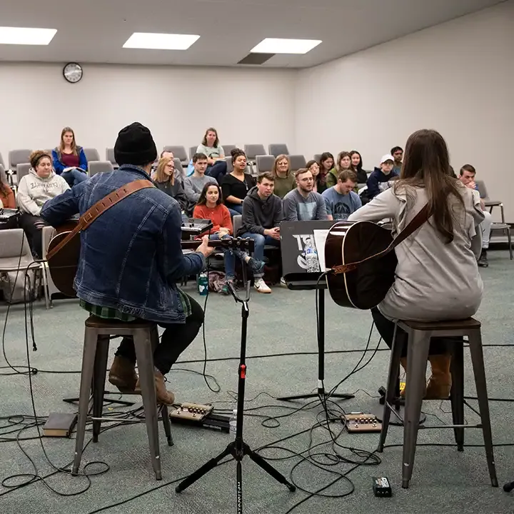 Two people playing guitars in room of people.